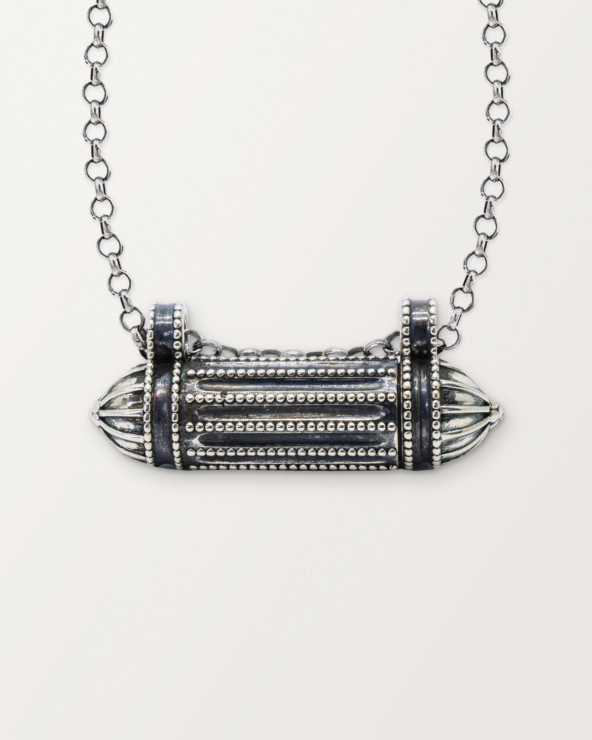 Close-up of the closed sterling silver Taweez amulet necklace, its beaded ribs and fluted end caps, on a warm bone-white background.