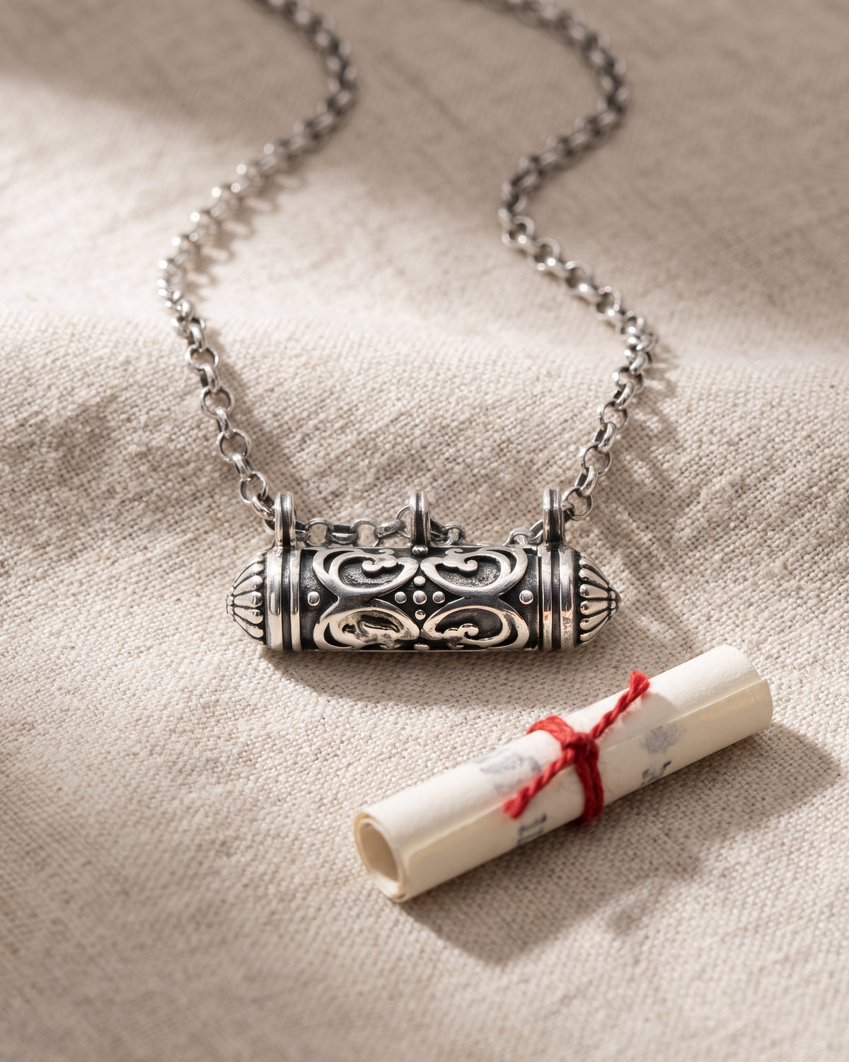 Illustrative still life: the Sri amulet necklace in sterling silver, closed, lying on oatmeal linen beside a small rolled paper scroll tied with red cotton thread.