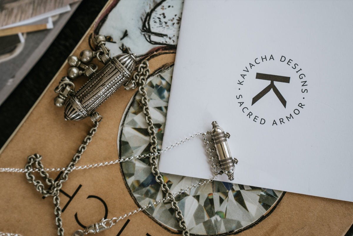 Two silver amulets on a sketchbook beside the Kavacha Designs seal.
