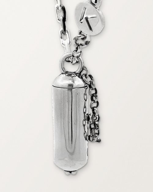 Close-up of the closed sterling silver Capsule Small amulet necklace, front, with its safety chain and K tag, on a warm bone-white background.