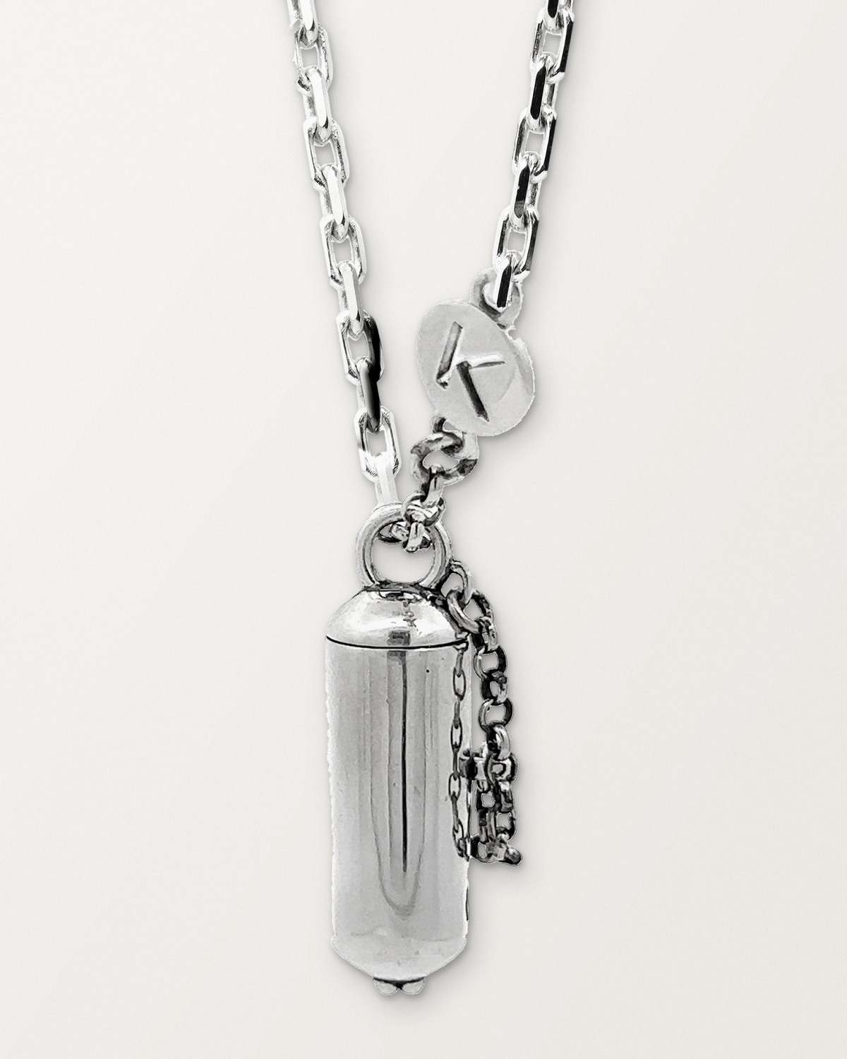 Sterling silver Capsule Small amulet necklace, closed, hanging on its chain with its safety chain and K tag, against a warm bone-white background.