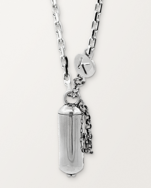 Sterling silver Capsule Small amulet necklace, closed, hanging on its chain with its safety chain and K tag, against a warm bone-white background.