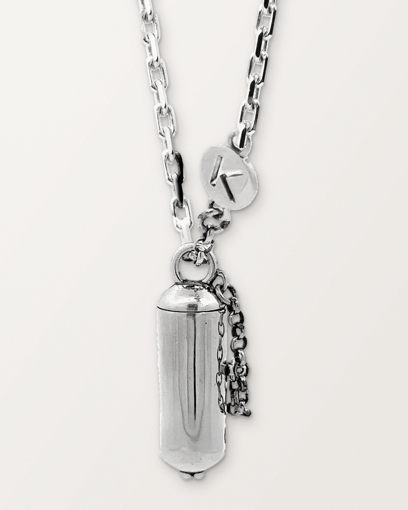 Sterling silver Capsule Small amulet necklace, closed, hanging on its chain with its safety chain and K tag, against a warm bone-white background.