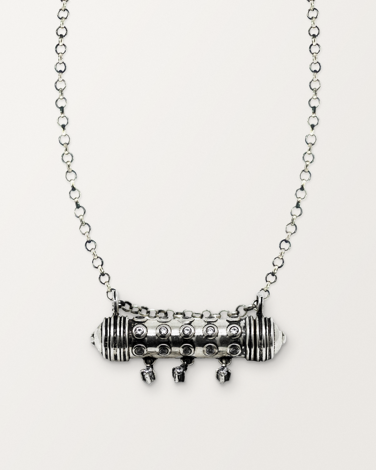Sterling silver Hirz amulet necklace, closed, with its three hanging stones, on a warm bone-white background, chain exiting the top of frame.