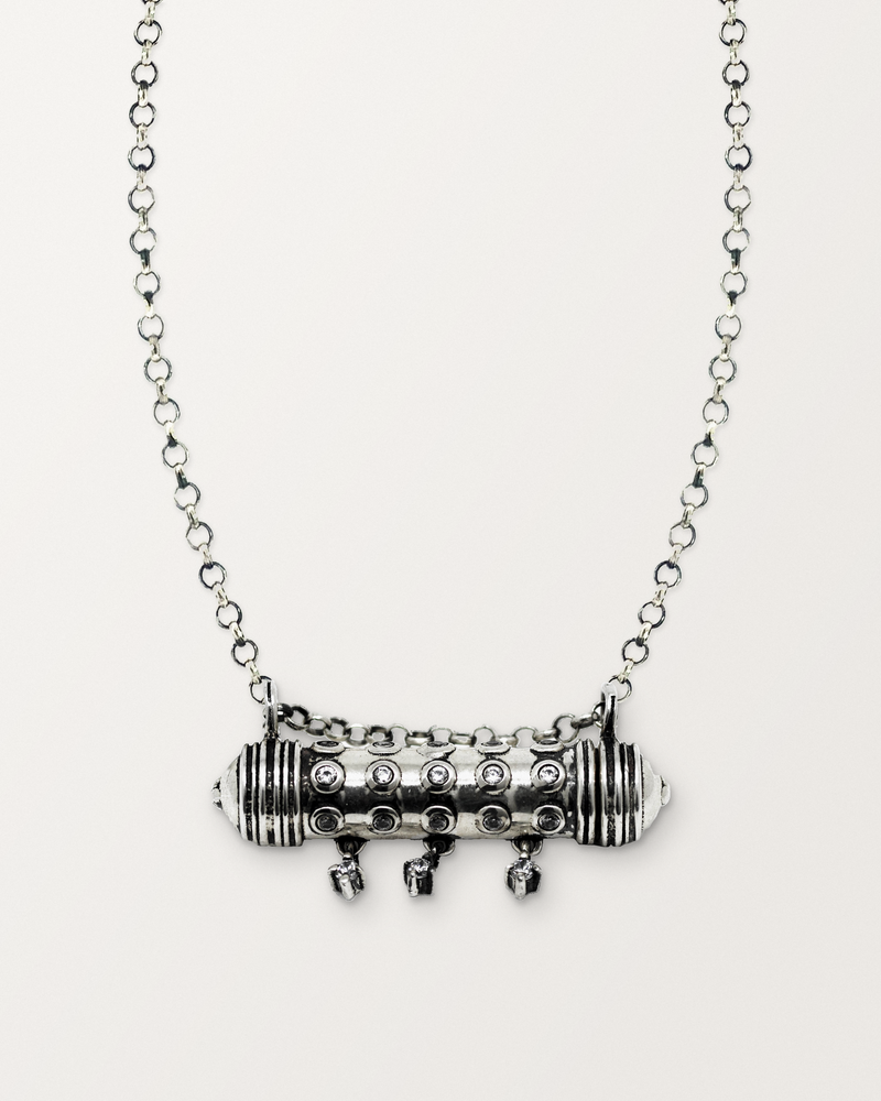 Sterling silver Hirz amulet necklace, closed, with its three hanging stones, on a warm bone-white background, chain exiting the top of frame.